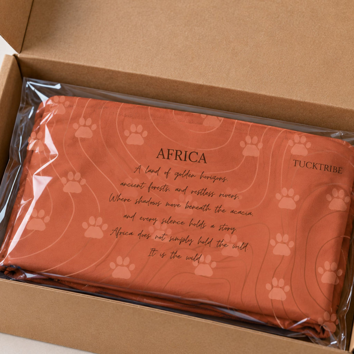 An terracotta orange neck gaiter with leopard paw prints and a printed poem titled 'AFRICA' by TUCKTRIBE, packaged in a clear plastic sleeve and placed inside a brown cardboard box.
