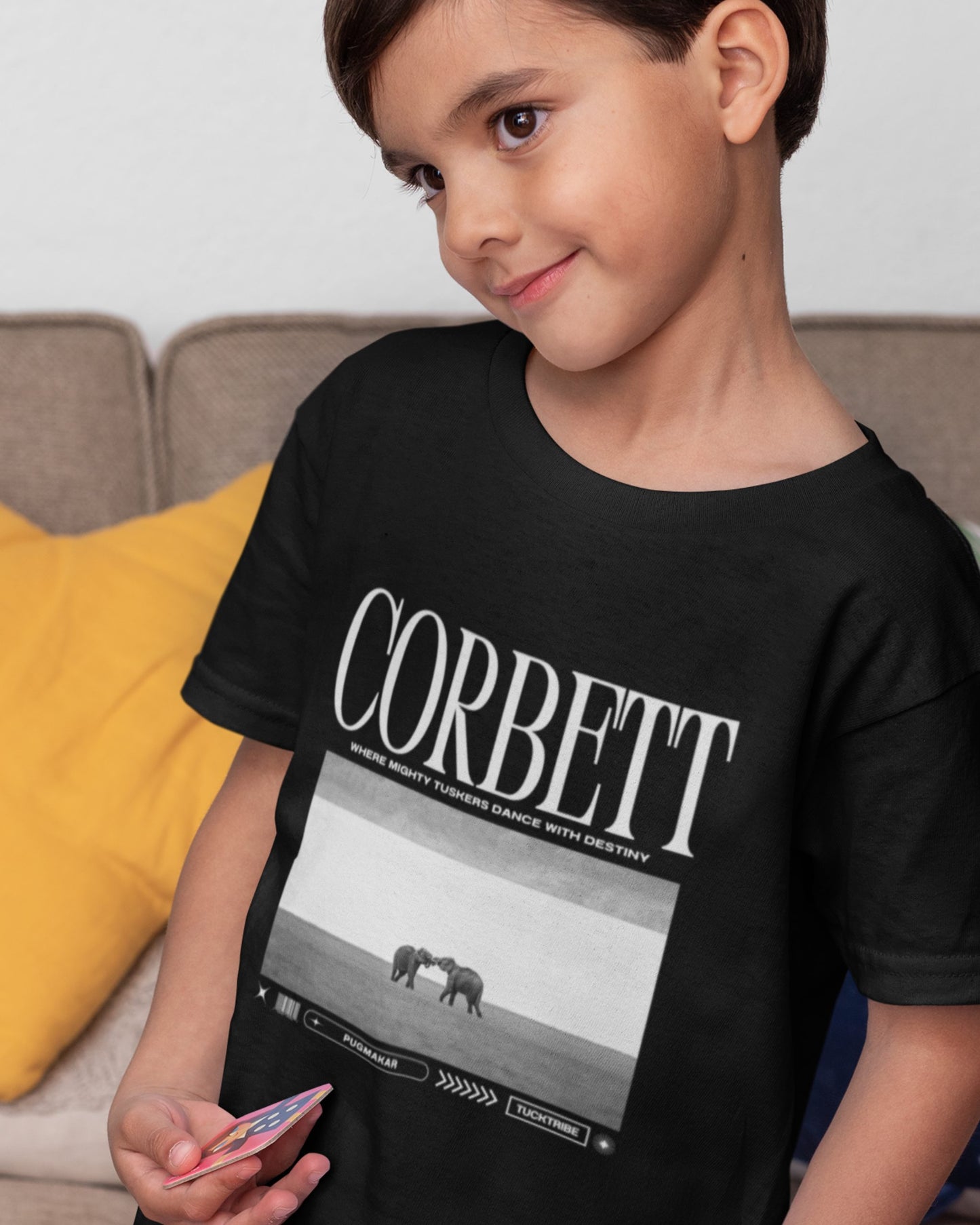 Child wearing a black t-shirt with 'Corbett' design, sitting on a couch.