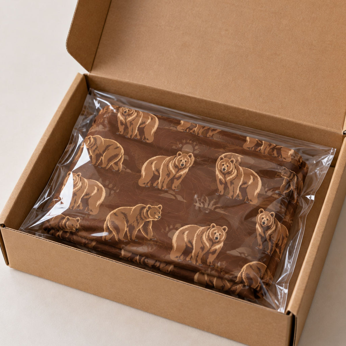 A brown fabric item with a repeating brown bear print, sealed in clear plastic packaging and displayed inside an open brown cardboard box on a light neutral surface.