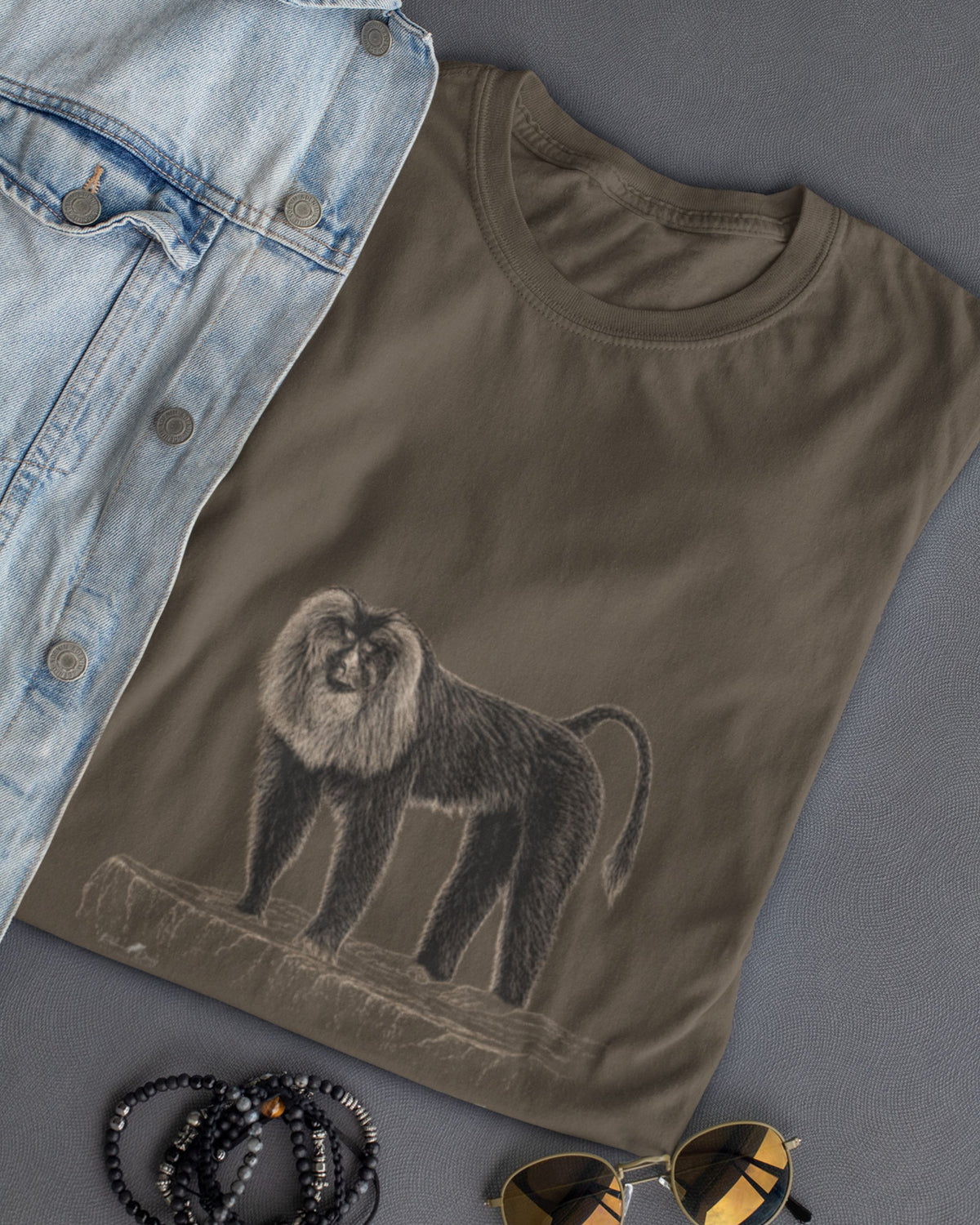 A folded olive green t-shirt featuring a detailed sketch of a lion tailed macaque styled in a flat-lay with a light blue denim jacket, black beaded bracelets, and gold-framed sunglasses on a gray background.