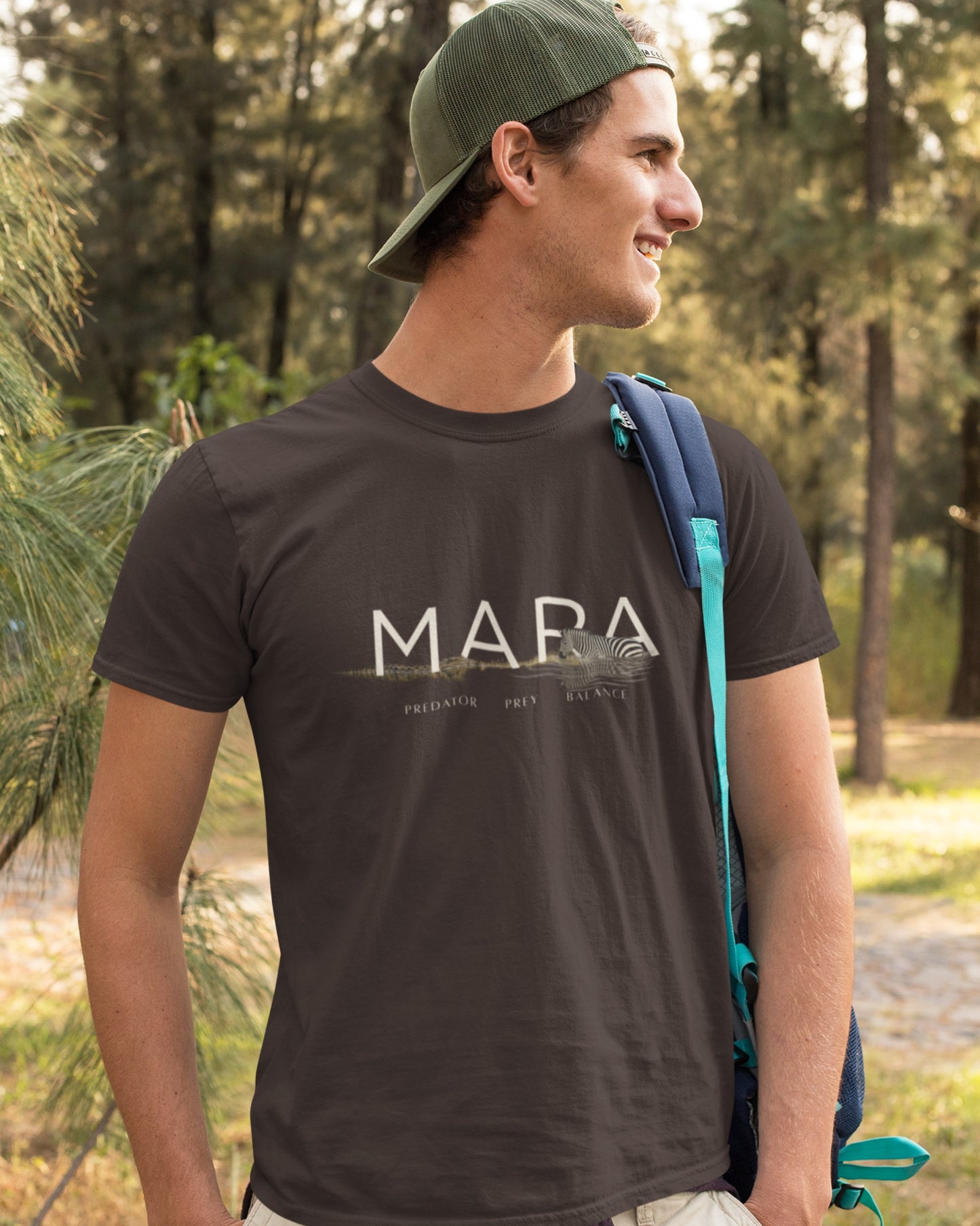 A man wearing a dark brown tucktribe t-shirt with a white 'MARA' graphic, a green cap, and a blue backpack, standing outdoors in a wooded area.