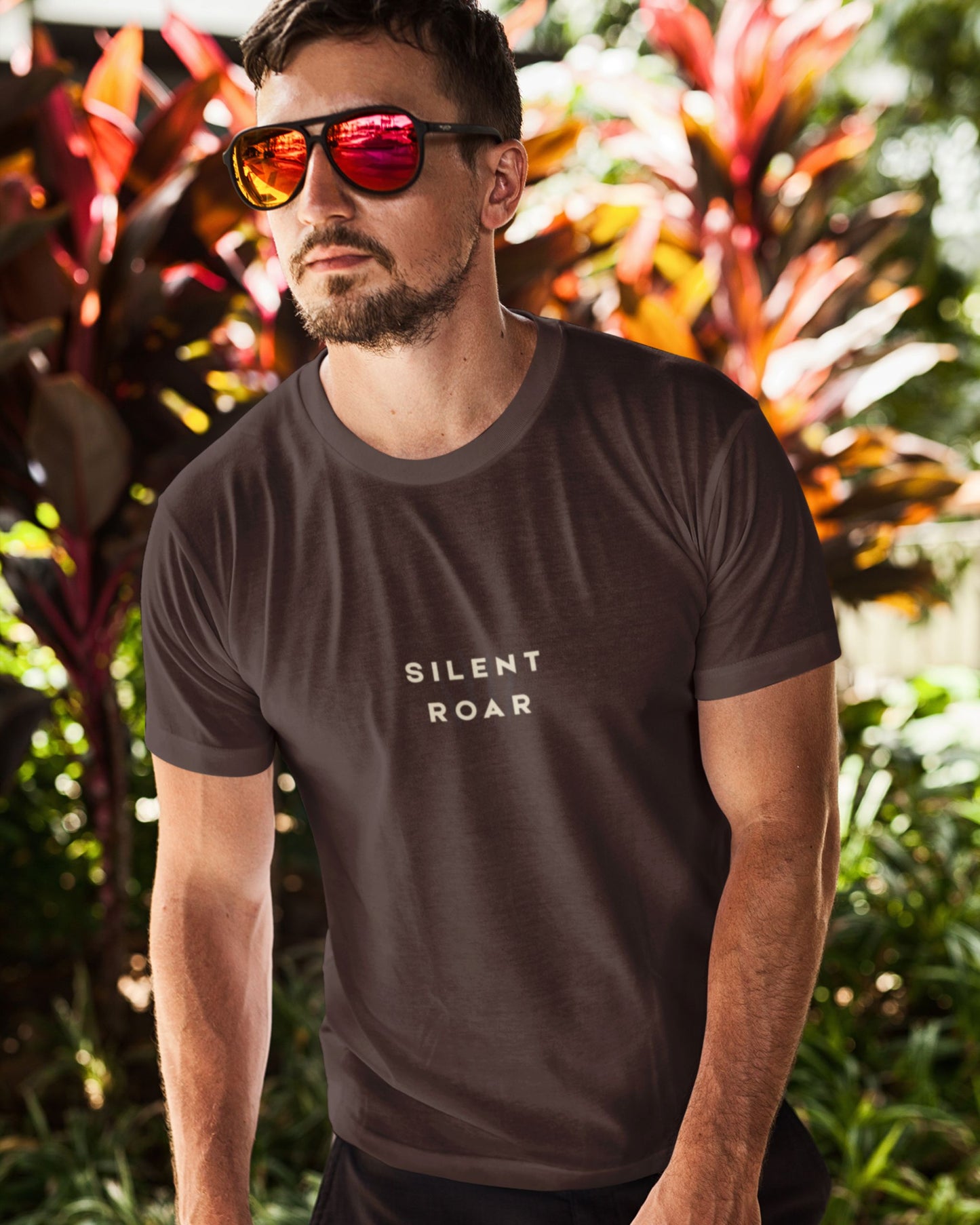 Man wearing a dark brown Tucktribe t-shirt with 'SILENT ROAR' text, standing in front of tropical plants.