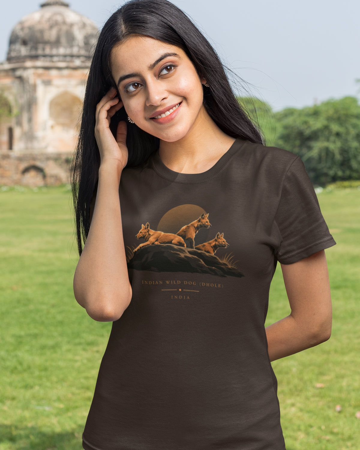 Woman wearing a brown Dhole wildlife T-shirt featuring three Indian wild dogs on a rocky landscape with sunset-inspired artwork