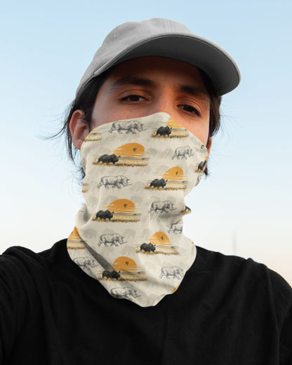 A person wearing a light gray baseball cap and a black shirt, with a beige neck gaiter featuring a safari print of rhinoceros and sunsets around their face, set against a pale blue sky.