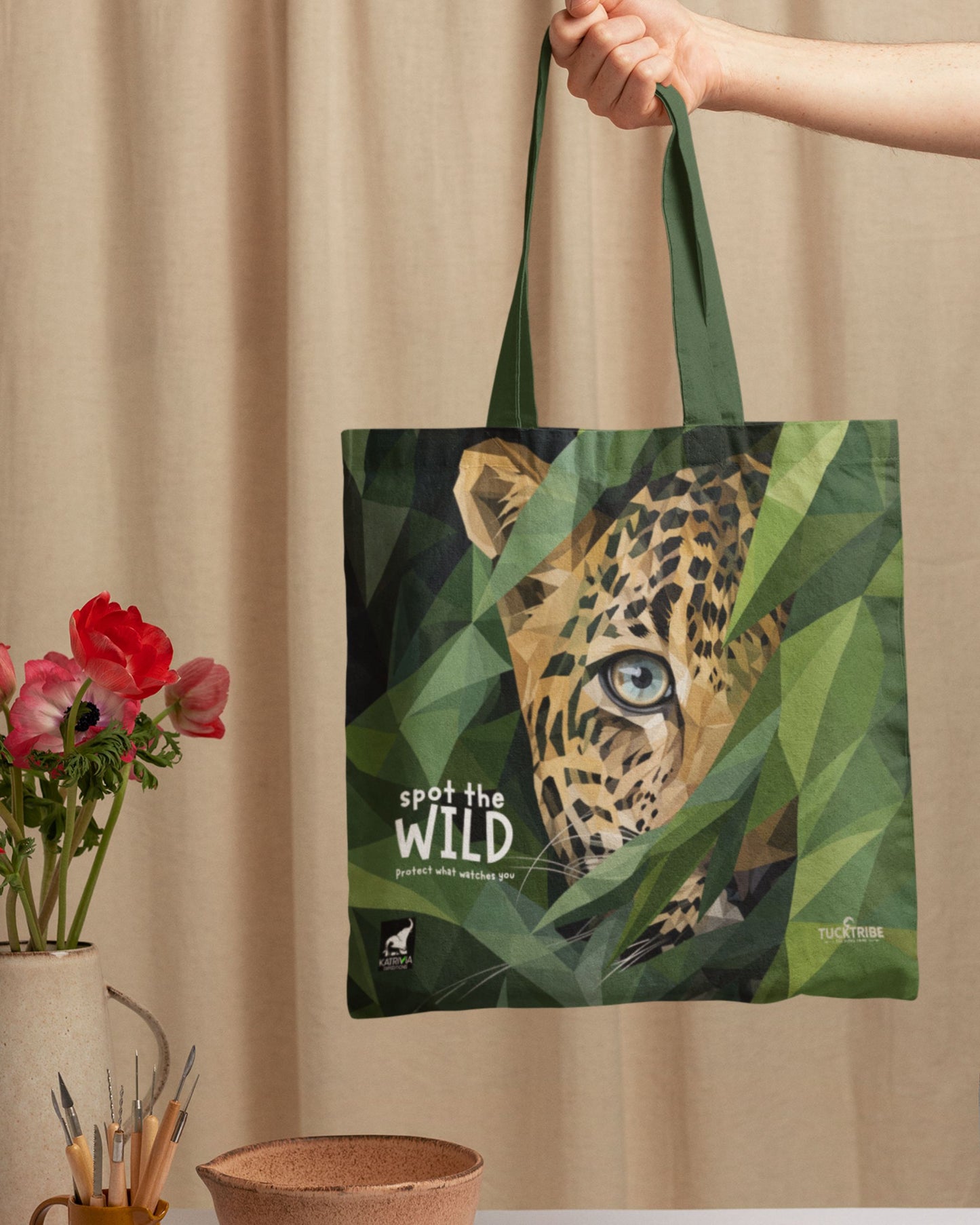 Tucktribe Tote bag with a leopard design and 'spot the wild' text, held by a hand against a beige curtain background.