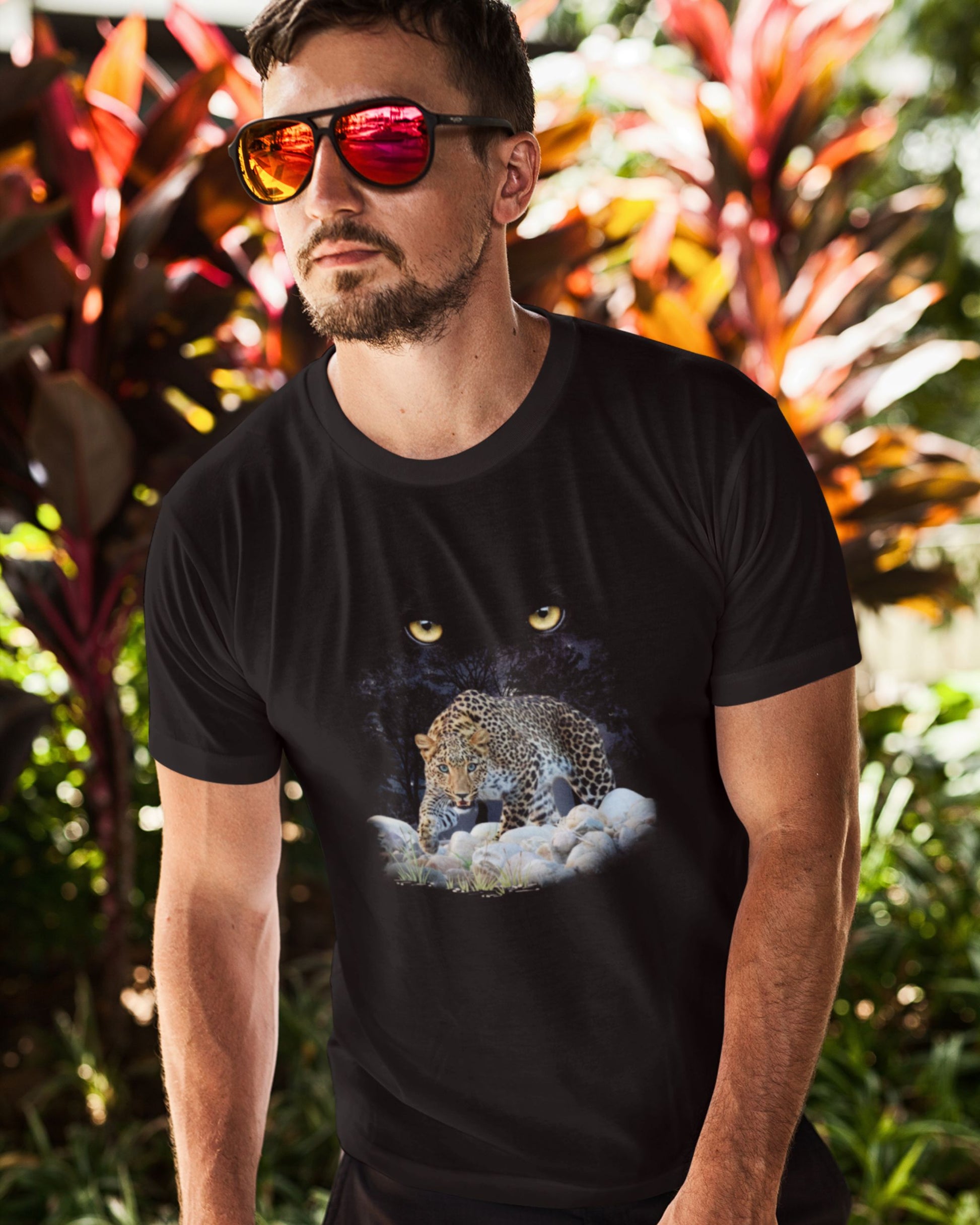 Man wearing a black tucktribe t-shirt with a graphic design of a leopard and clouds, standing in front of tropical plants.