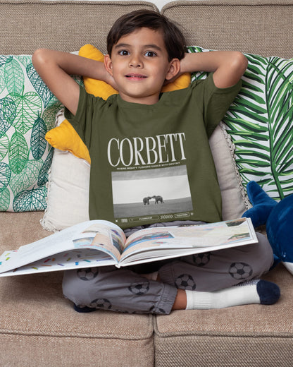 Child wearing a green t-shirt with 'Corbett' printed on it, sitting on a couch with an open book.