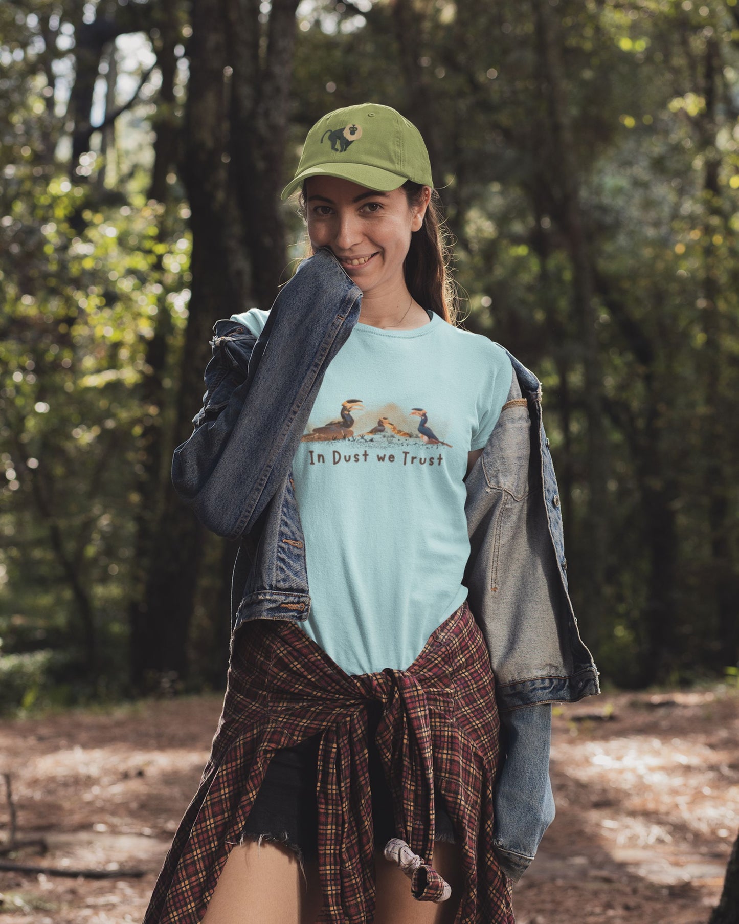 Woman in a forest wearing a mint colour t-shirt with text and graphics, a green cap, and a plaid skirt.