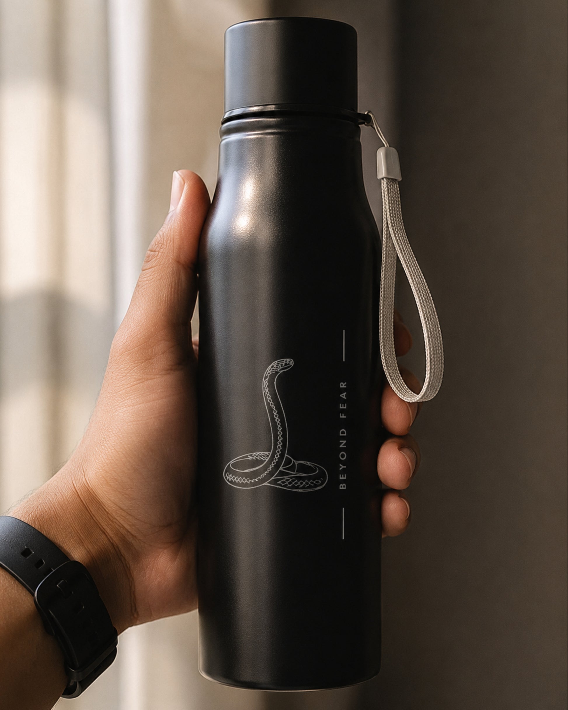closeup of a matte black insulated tucktribe water bottle held in hand with a matching black screw cap and a white wrist strap, featuring a engraving of a cobra and the text 'BEYOND FEAR' on the front, set against a warm interior.