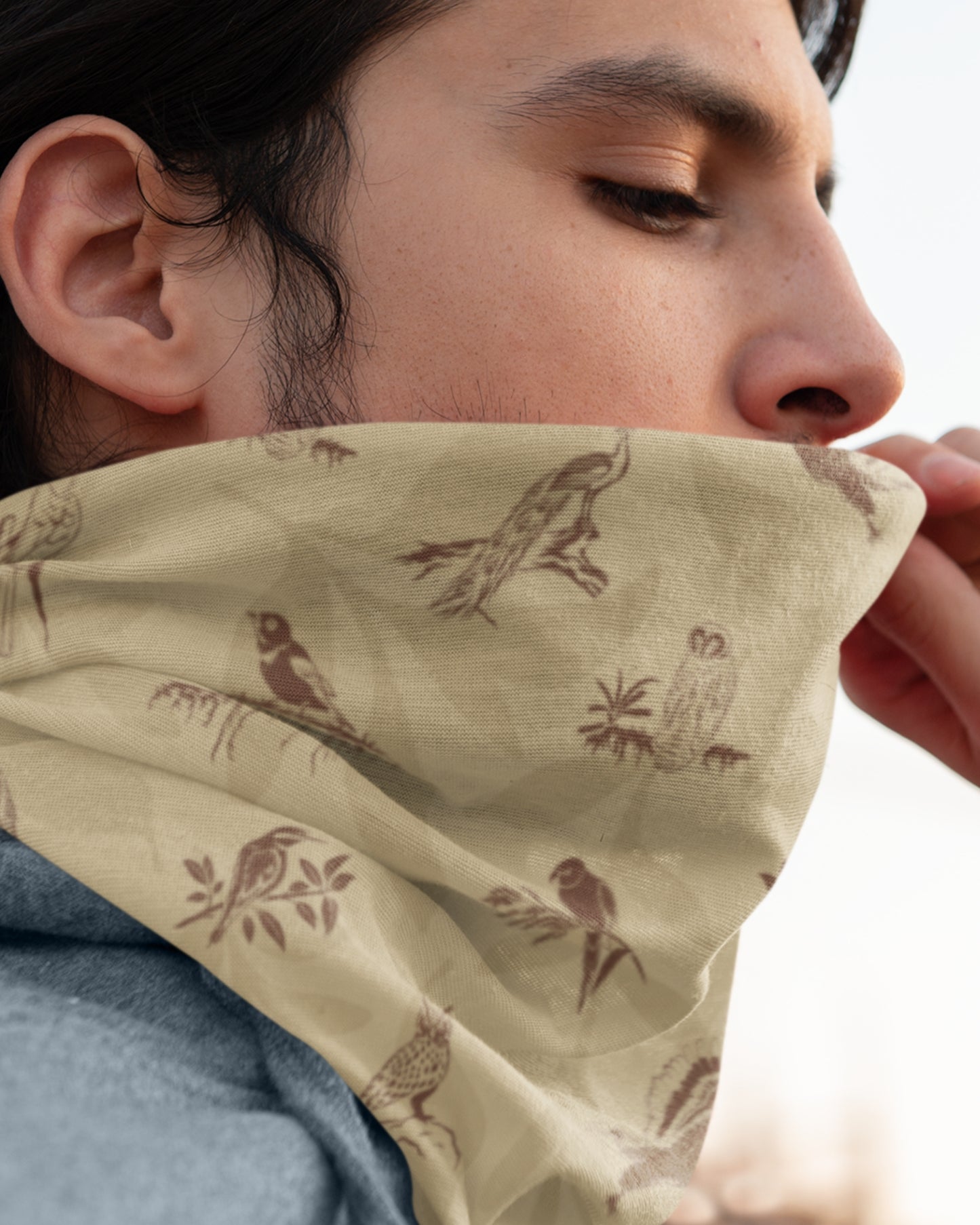 A close-up lifestyle shot of a person wearing a beige neck gaiter with a brown bird and owl pattern, pulled up over the lower face.