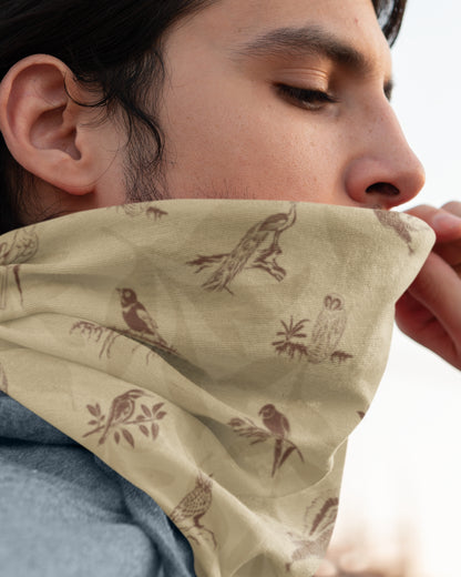 A close-up lifestyle shot of a person wearing a beige neck gaiter with a brown bird and owl pattern, pulled up over the lower face.