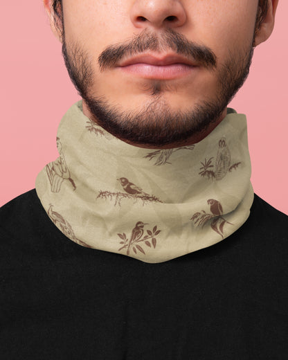 A close-up of a man wearing a light beige neck gaiter with a brown bird and branch pattern, paired with a black shirt against a solid pink background.