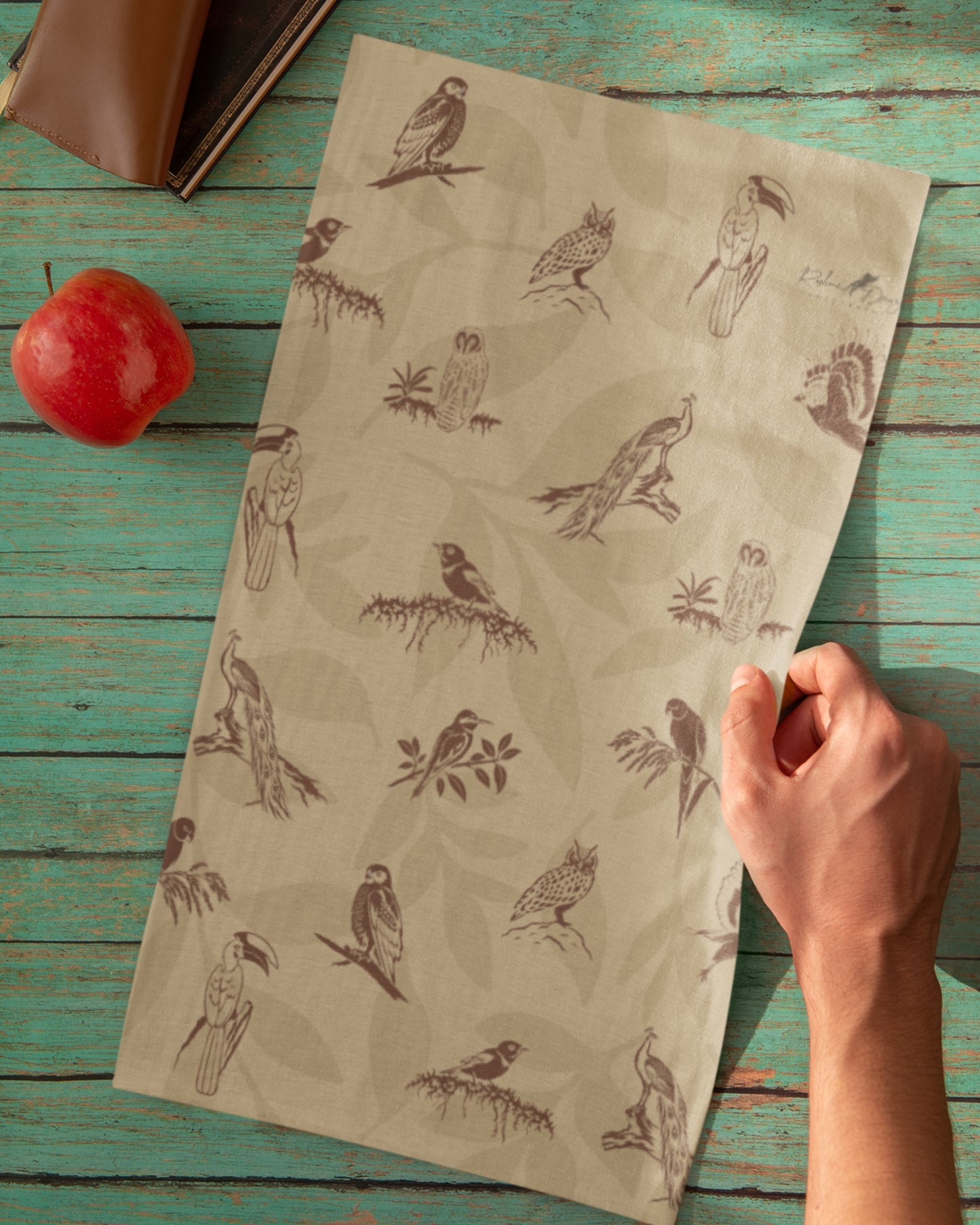 A beige neck gaiter with a brown bird and owl pattern, held by a hand over a rustic teal wooden table with a red apple and a book as props.