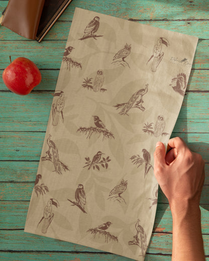A beige neck gaiter with a brown bird and owl pattern, held by a hand over a rustic teal wooden table with a red apple and a book as props.
