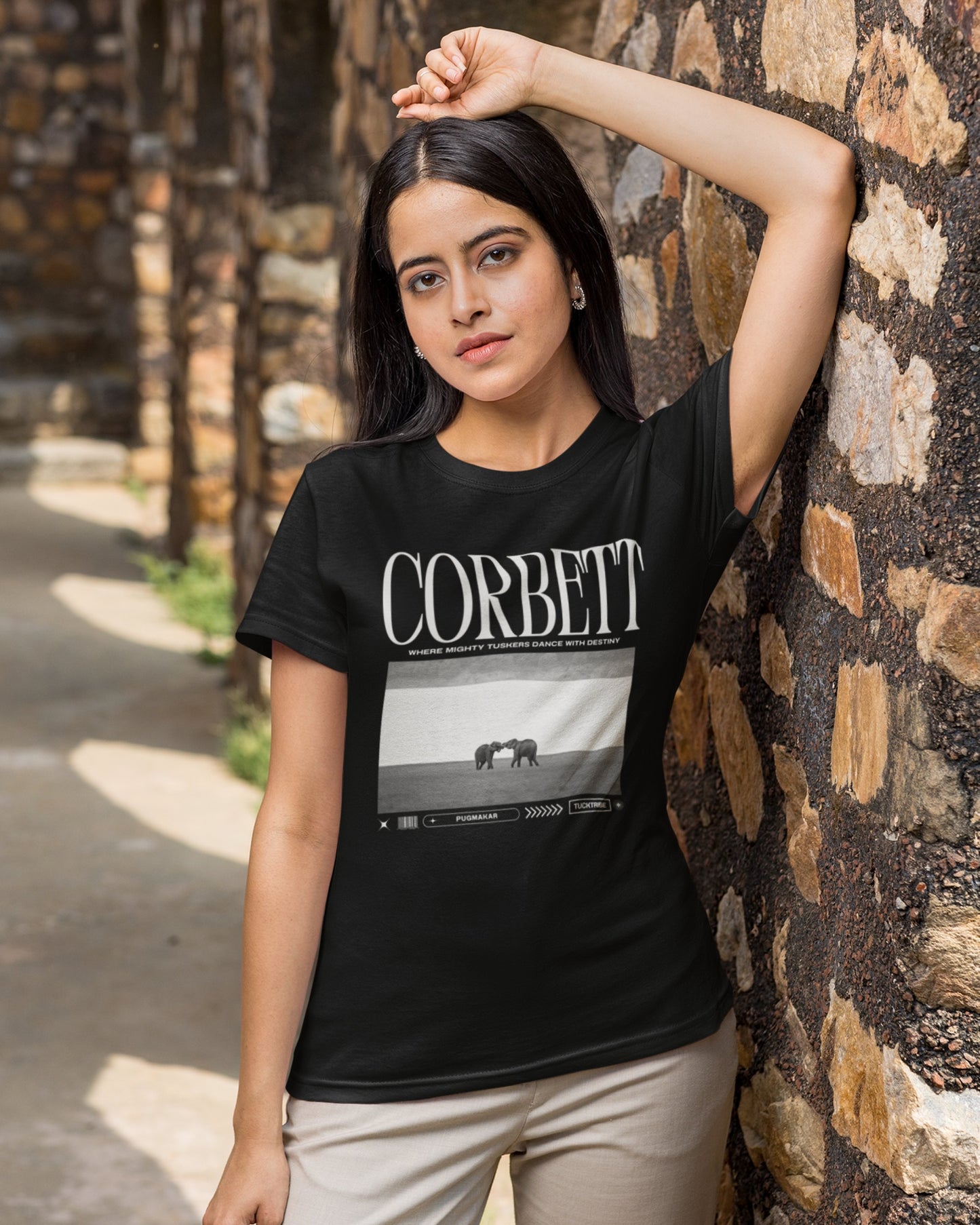 Woman wearing a black t-shirt with 'Corbett' graphic design against a stone wall.