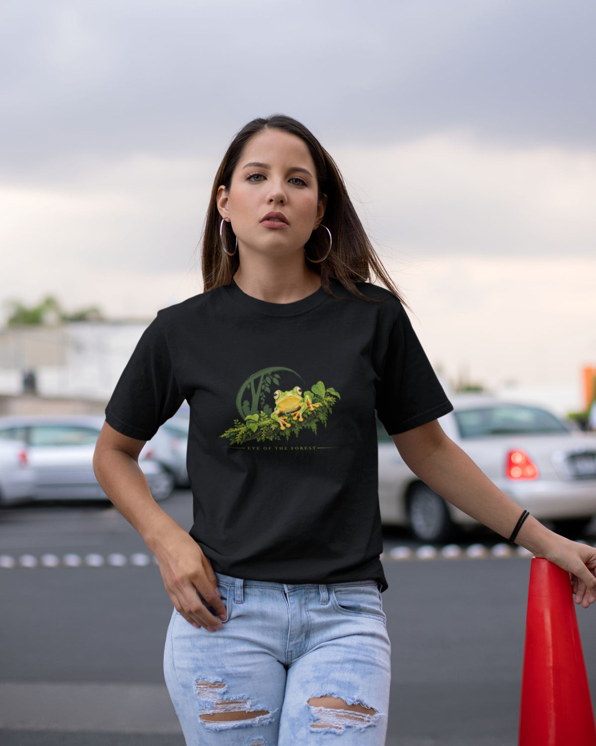 A woman wearing a black graphic t-shirt with a malabar gliding frog design, paired with light-wash distressed jeans, standing in an outdoor parking lot.