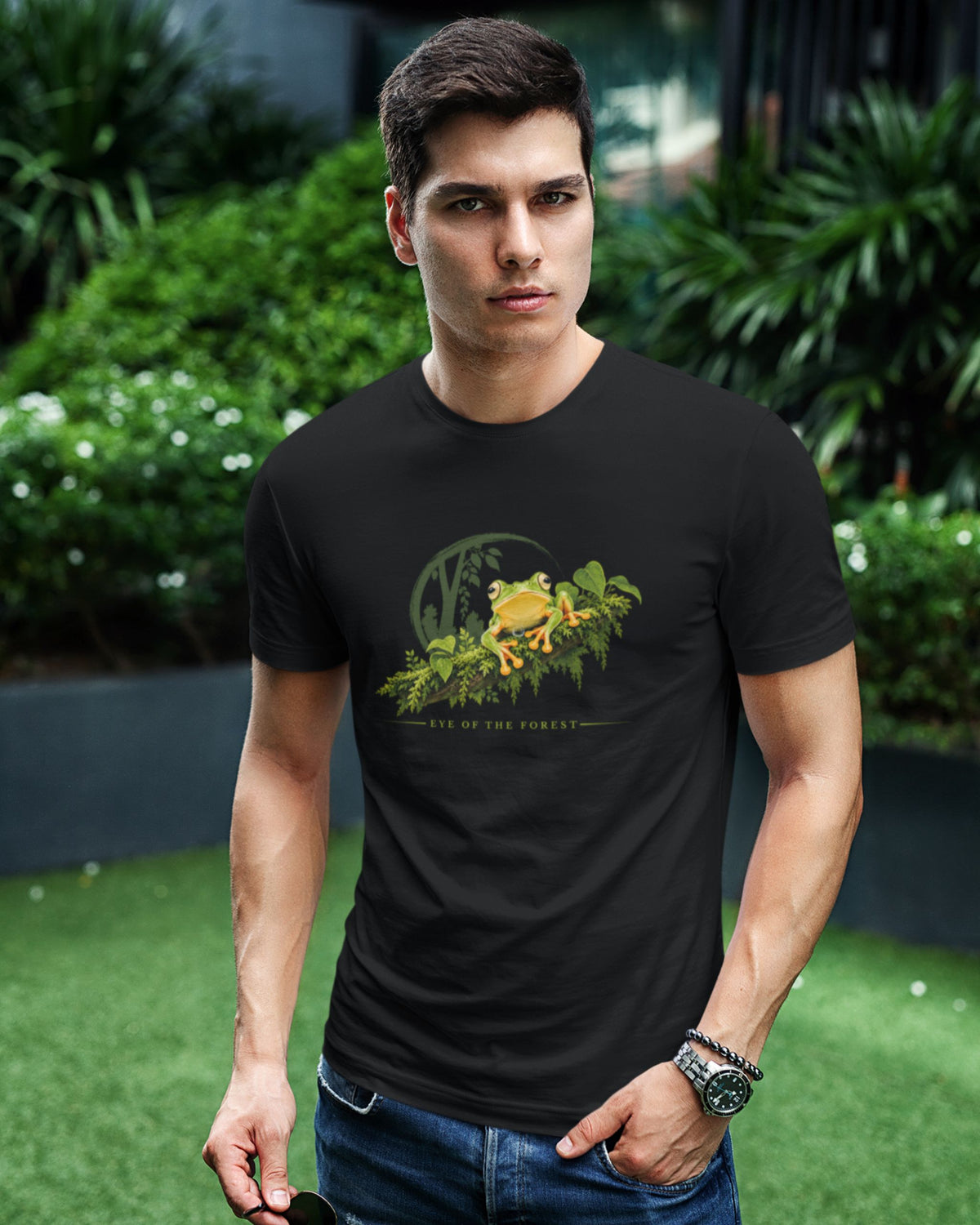 A man wearing a black short-sleeve t-shirt with a green and yellow malabar gliding frog graphic on the chest, standing outdoors in front of a blurred green garden background.