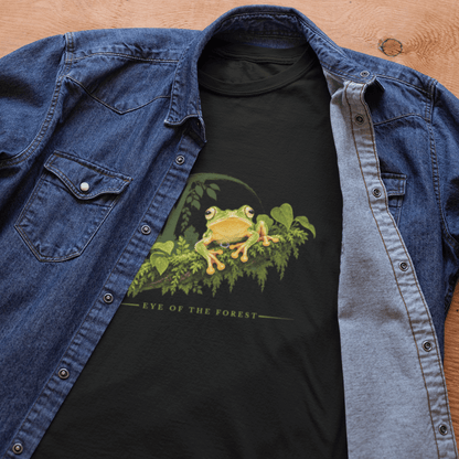 A black graphic t-shirt featuring a green and yellow malabar gliding frog on the chest, styled under an open blue denim shirt on a wooden background. The text 'EYE OF THE FOREST' is printed below the graphic.