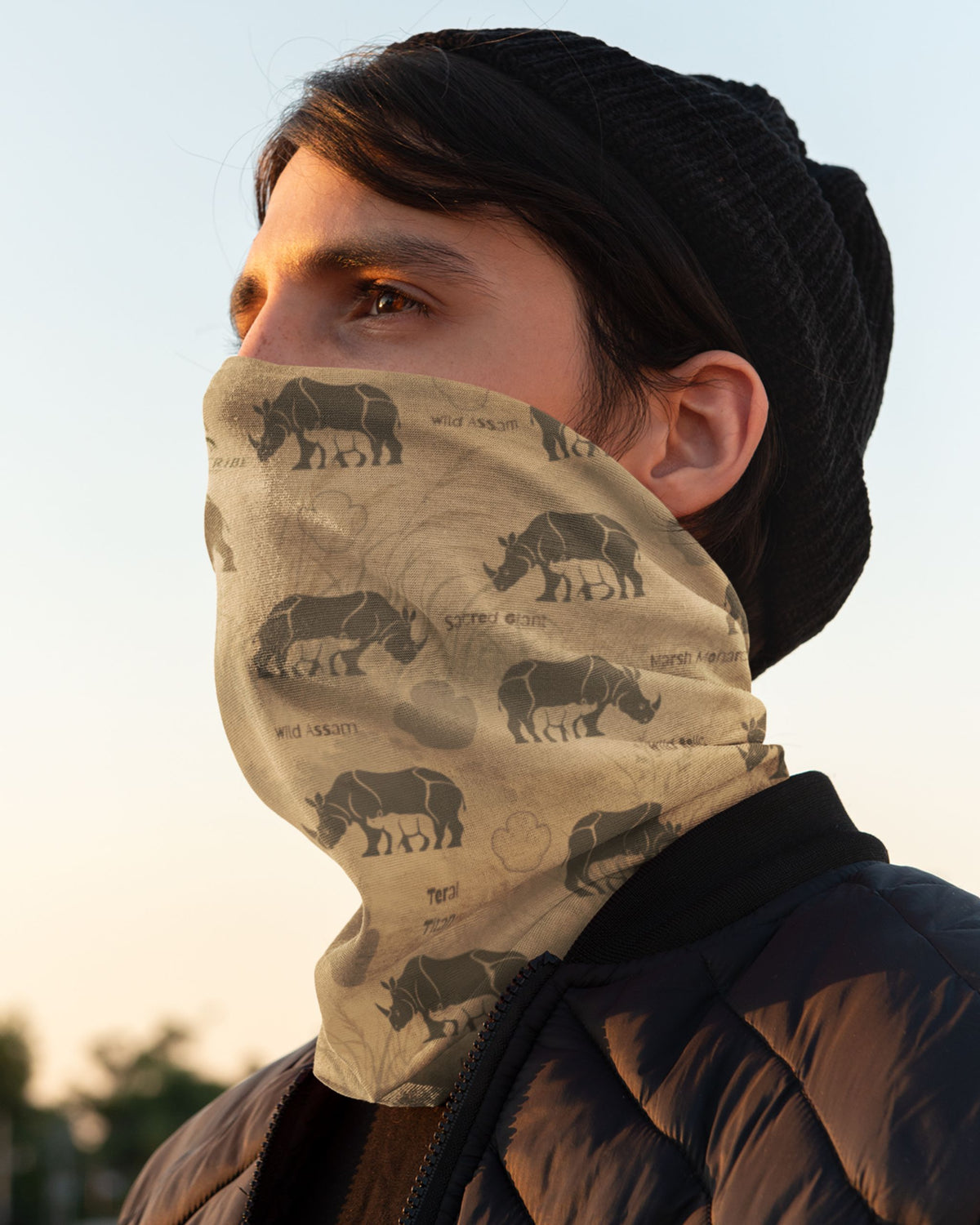 A person wearing a beige neck gaiter with a black rhinoceros print, paired with a black beanie and a dark puffer jacket, shown in profile against a soft sky background.
