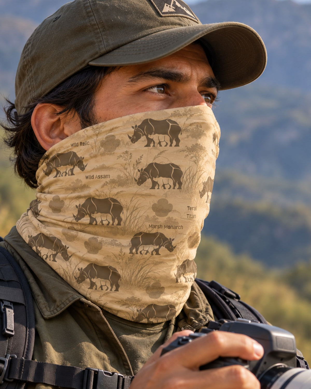 A close-up lifestyle shot of a man wearing a beige neck gaiter with a rhinoceros print, paired with an olive green cap and outdoor gear, with a blurred mountain background.