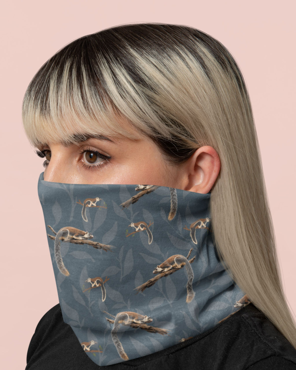A close-up of a woman wearing a blue-gray neck gaiter with a printed pattern of grizzled giant squirrel and leaves, set against a solid pale pink background.