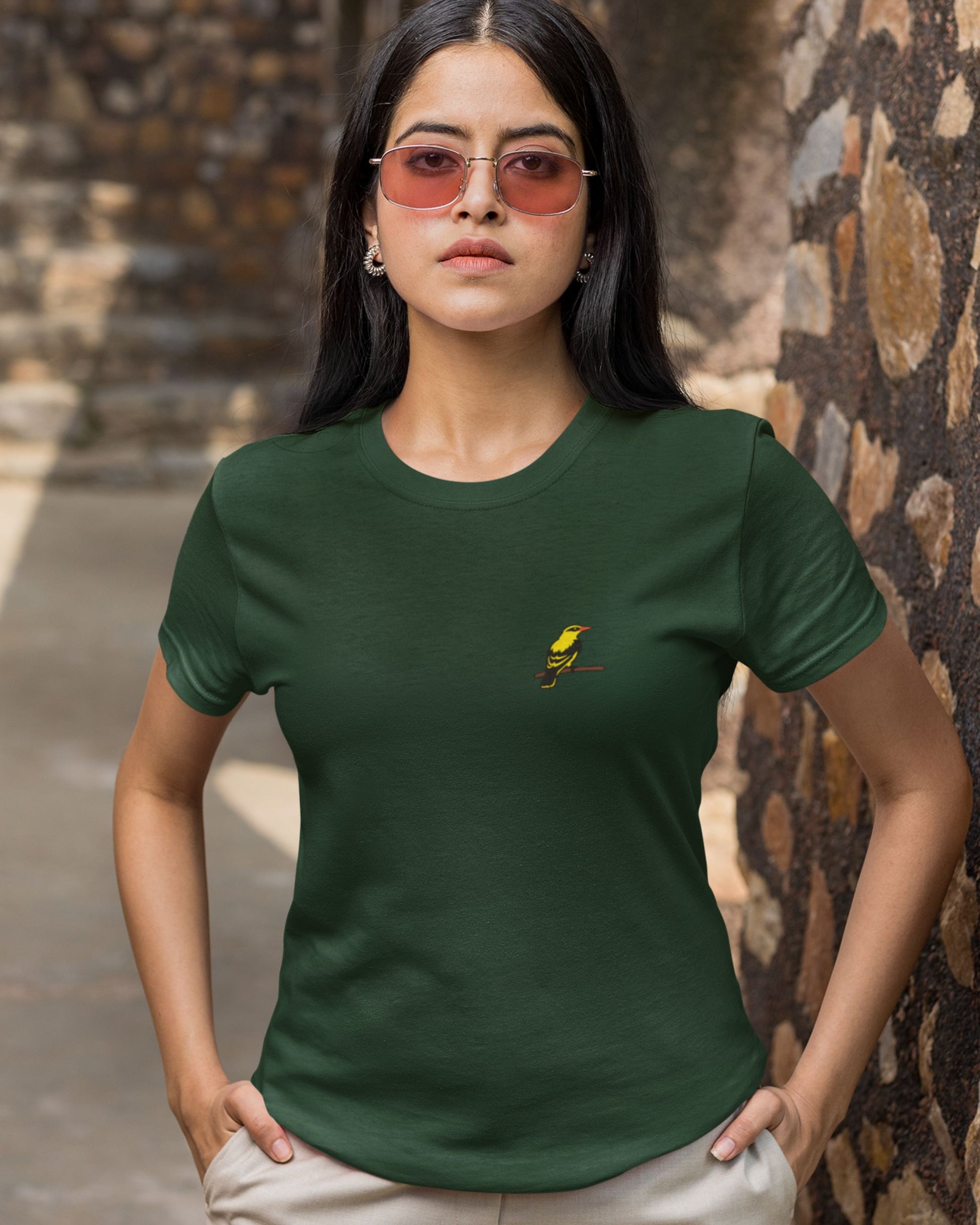 Woman wearing a Tucktribe green t-shirt with a embroidery of a golden oriole bird against a stone wall background