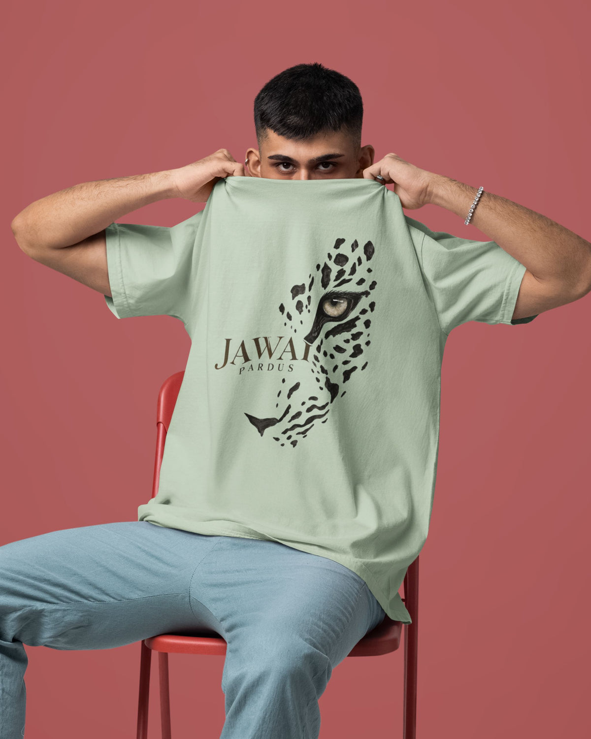 A man wearing a sage green t-shirt with a black leopard graphic and 'JAWAI PARDUS' text, holding the shirt up to show the front print. He is sitting on a red chair against a solid dusty rose background.