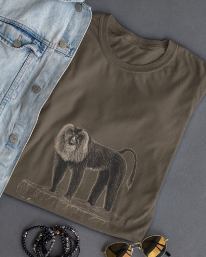 A folded olive green t-shirt featuring a detailed sketch of a lion tailed macaque  styled in a flat-lay with a light blue denim jacket, black beaded bracelets, and gold-framed sunglasses on a gray background.