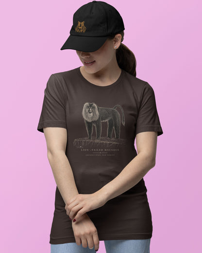 A woman wearing a dark brown t-shirt with a detailed illustration of a lion tailed macaque on the front, paired with a black baseball cap and light blue jeans, set against a solid pink background.