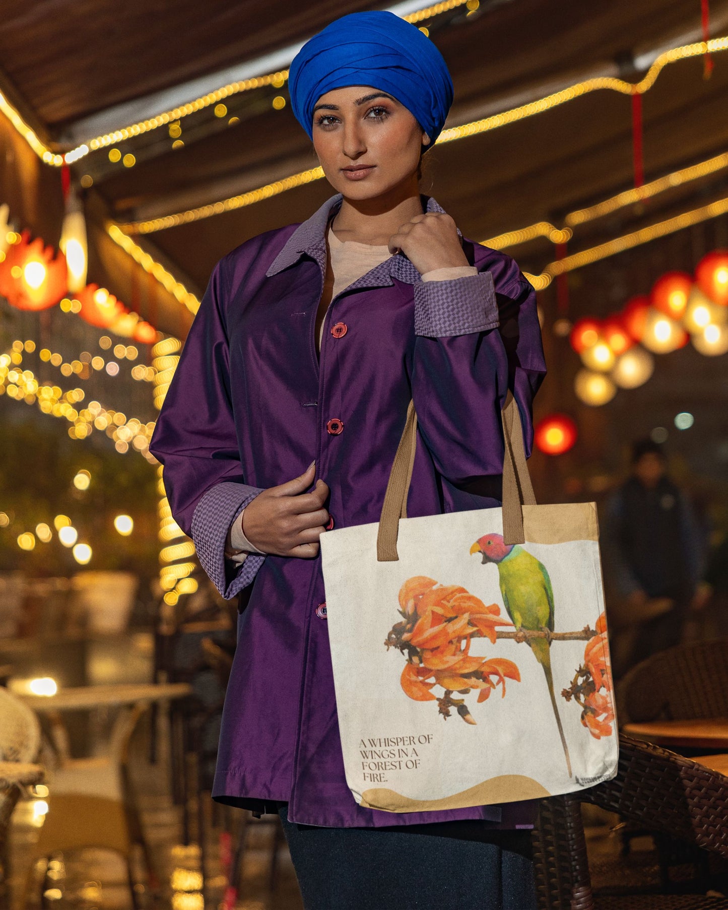Person wearing a purple coat and blue turban holding a tucktribe tote bag with a plum headed parakeet design in an outdoor setting.