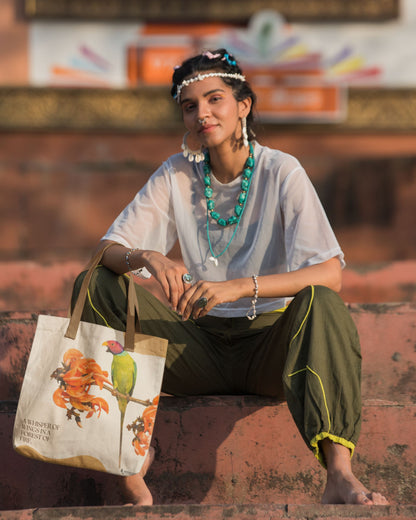 Woman sitting on steps holding a tucktribe tote bag with a plum headed parakeet design, wearing traditional attire.