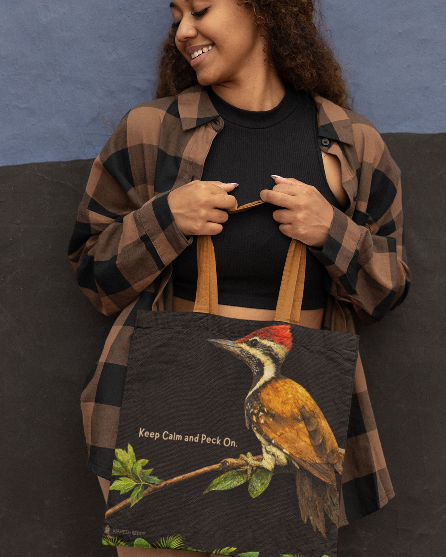 Woman holding a tucktribe tote bag with a woodpecker design and text, against a blue wall.