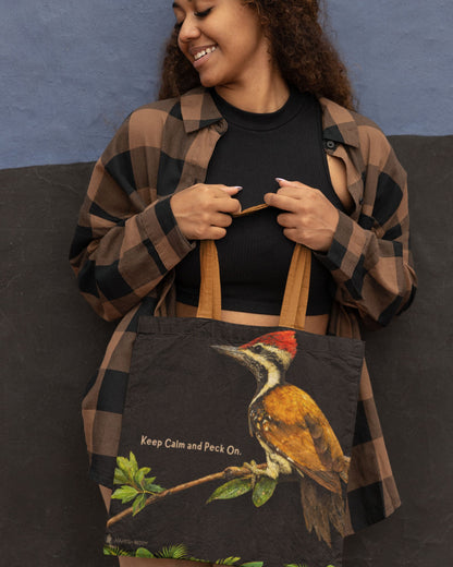 Woman holding a tucktribe tote bag with a woodpecker design and text, against a blue wall.
