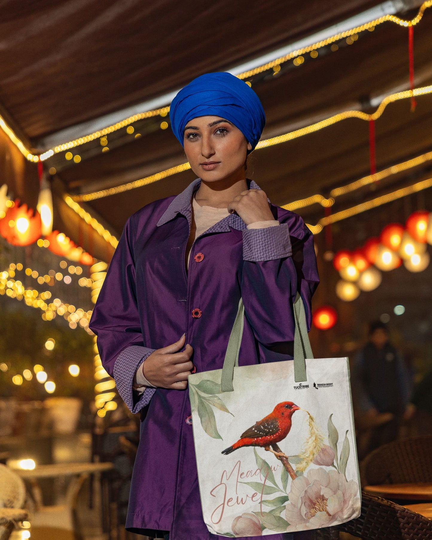 Woman in a purple coat and blue turban holding a tucktribe tote bag with a red munia bird design, outdoor setting with lights.