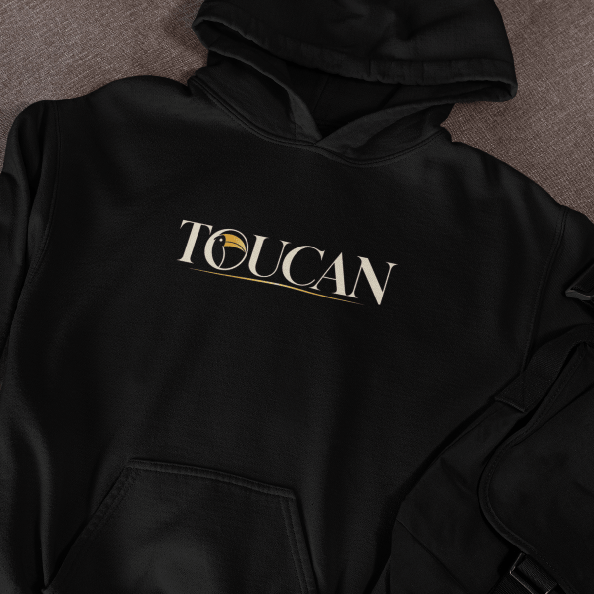 A black pullover hoodie laid flat on a textured gray surface, featuring the word 'TOUCAN' printed across the chest in white with a gold circular emblem and underline detail.