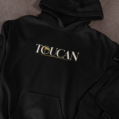 A black pullover hoodie laid flat on a textured gray surface, featuring the word 'TOUCAN' printed across the chest in white with a gold circular emblem and underline detail.