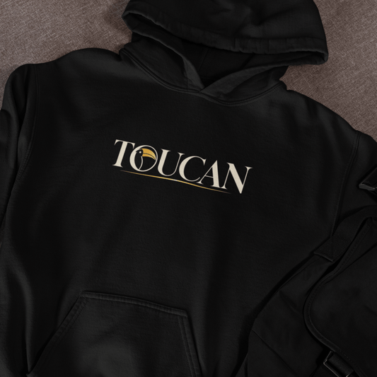 A black pullover hoodie laid flat on a textured gray surface, featuring the word 'TOUCAN' printed across the chest in white with a gold circular emblem and underline detail.