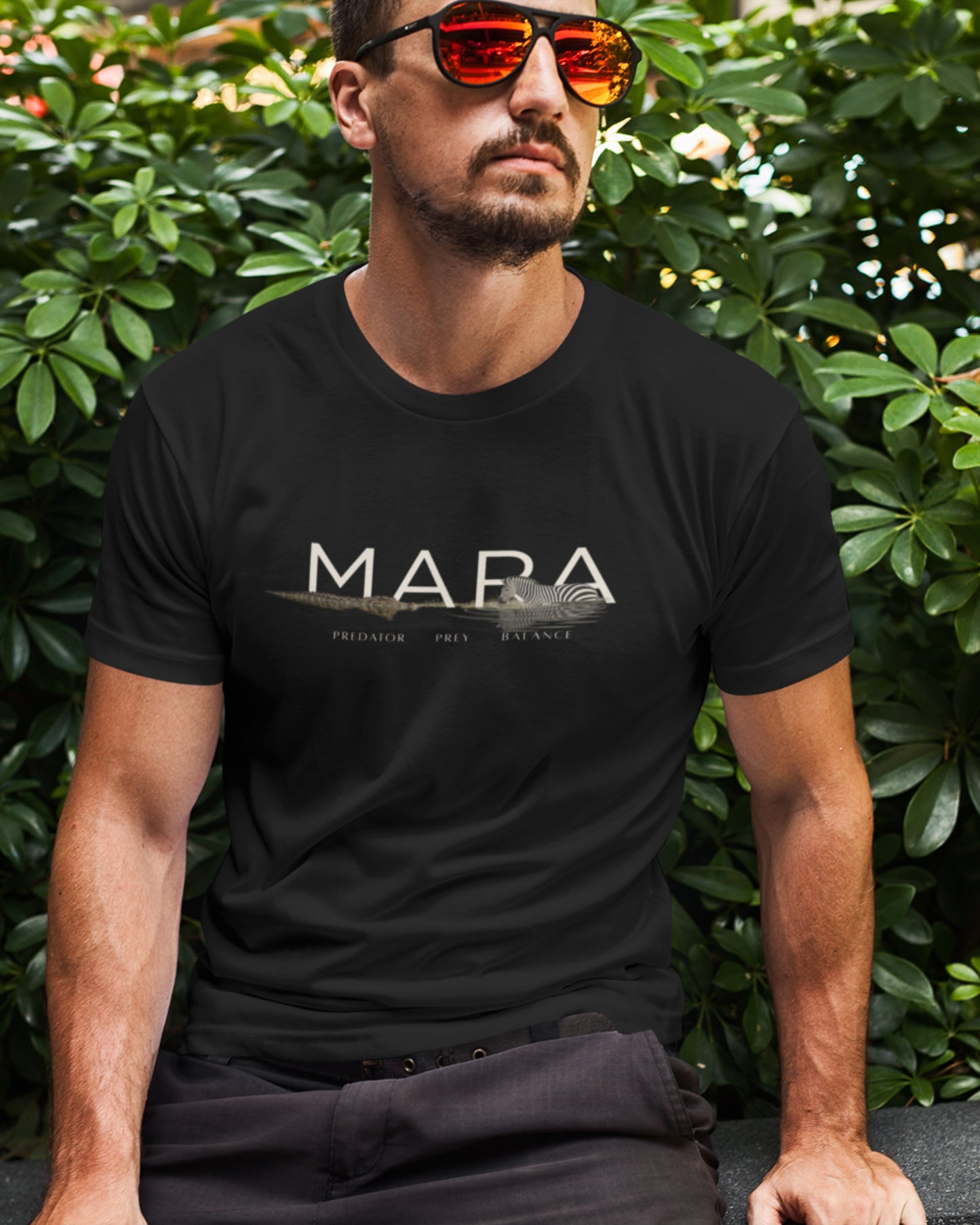 A man wearing a black tucktribe crewneck t-shirt with a white 'MARA' graphic and a small sailboat illustration across the chest, set against a background of green foliage.