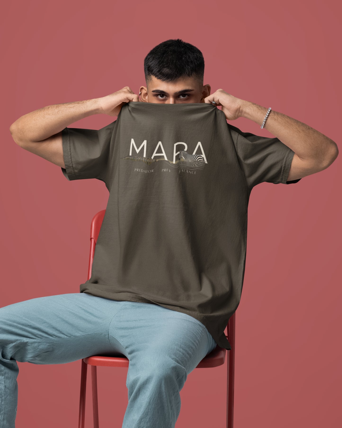 A man wearing  a olive green Tucktribe short-sleeve crewneck t-shirt with a beige graphic print on the chest featuring the word 'MARA' and smaller text 'PREDATOR - PREY- BALANCE' with a Zebra and crocodile illustration, displayed against a solid Orange background.