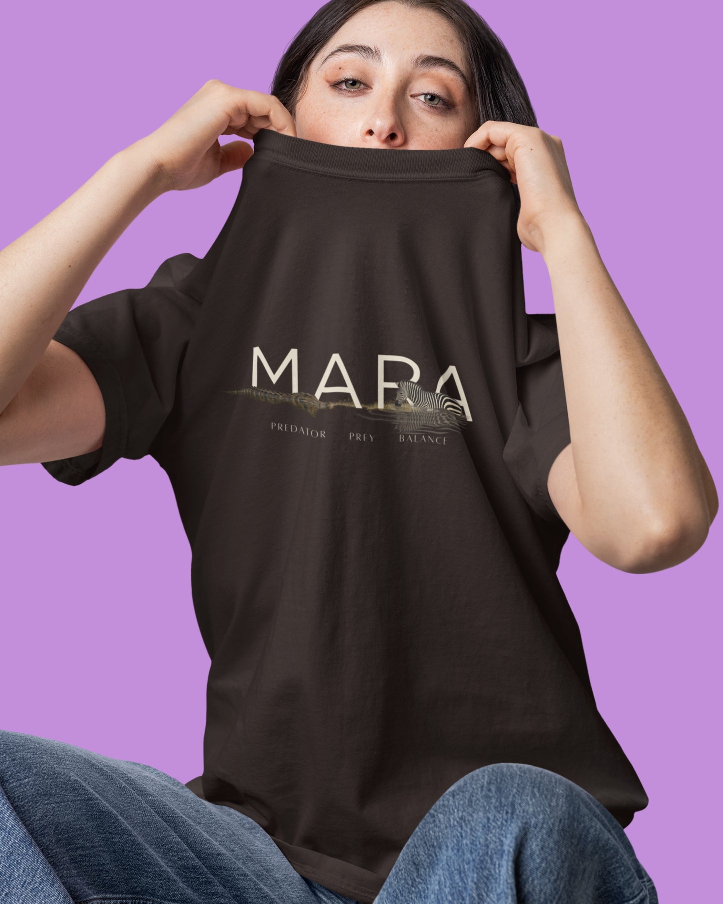 A woman holding up a dark brown tucktribe t-shirt with 'MARA' printed in white across the chest, set against a solid purple background.