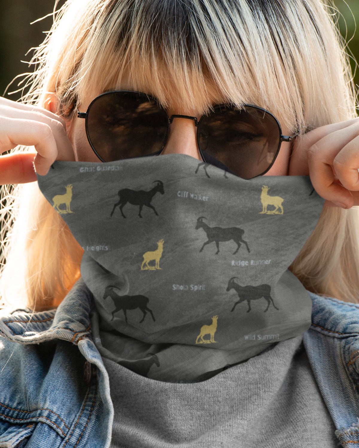 A close-up lifestyle shot of a person wearing a dark gray neck gaiter with a repeating pattern of black and yellow Nilgiri Thar and text labels, styled with sunglasses and a denim jacket.