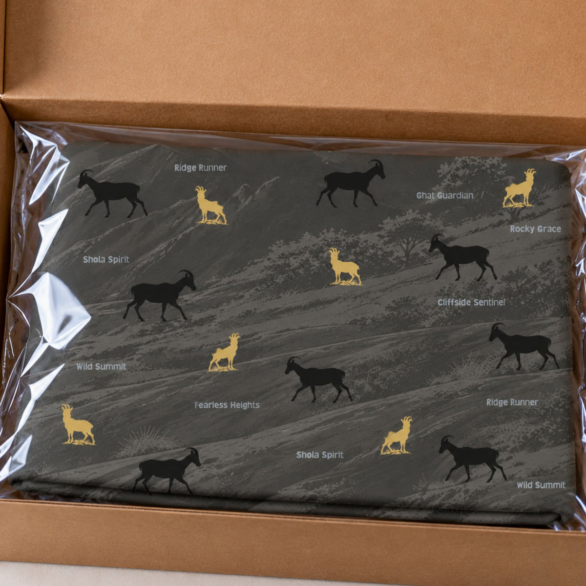 A folded dark gray neck gaiter, wrapped in clear plastic and placed inside an open brown cardboard box. The fabric features a pattern of black and yellow nilgiri thar silhouettes with text printed next to each one.