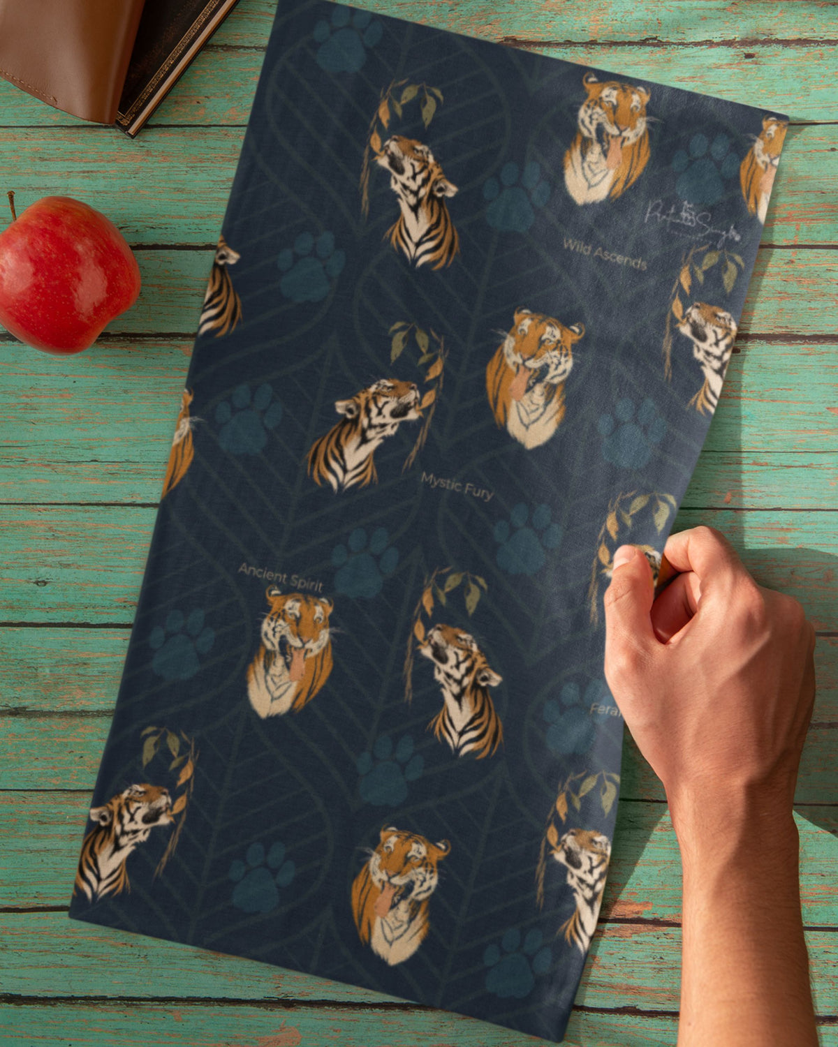 A dark teal neck gaiter, featuring a pattern of tiger faces and paw prints on a dark navy blue background, held by a hand over a rustic green wooden table with a red apple and a brown leather item as props.