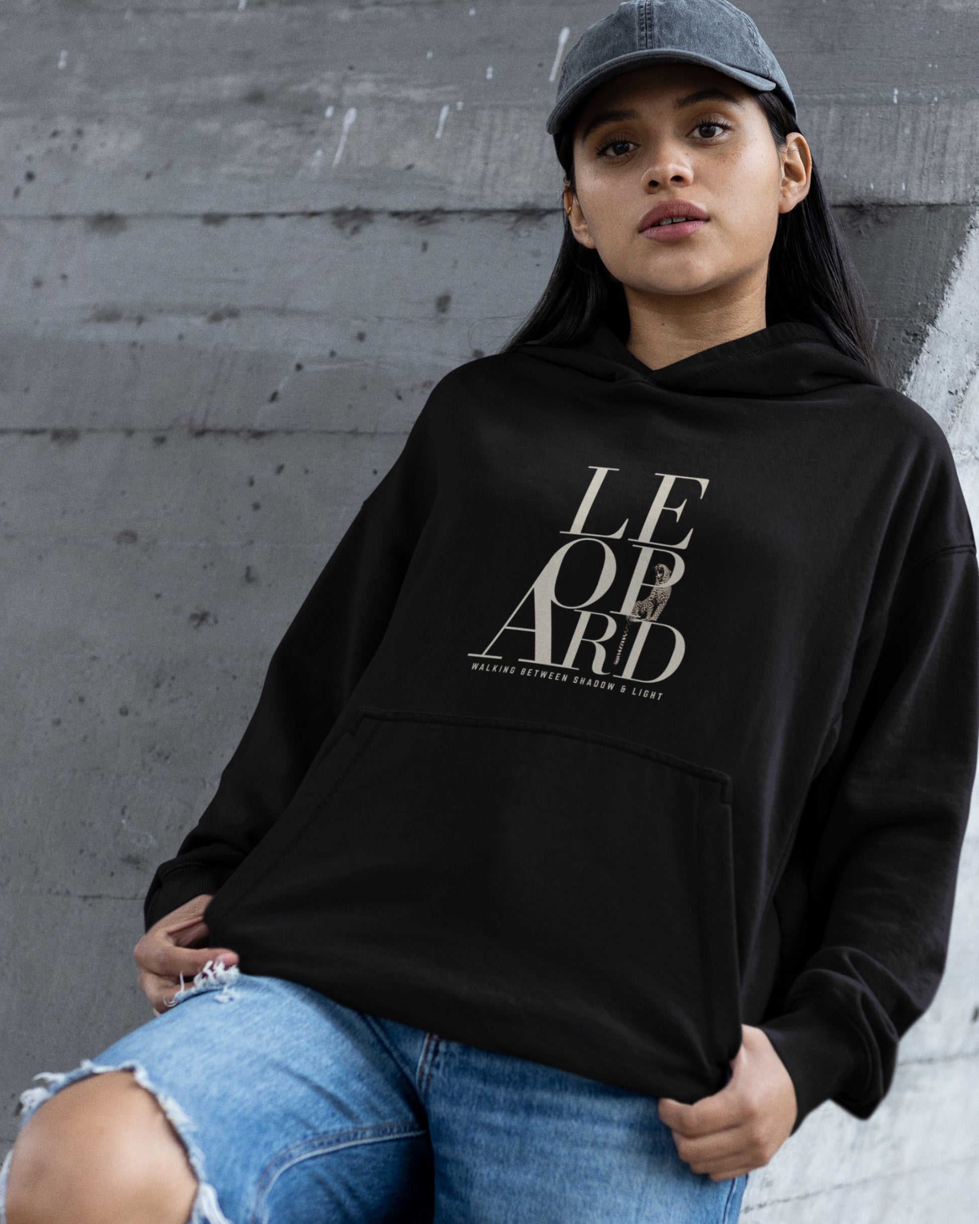 A woman wearing a black pullover hoodie with a large leopard Text and Walking Between Shadow and Light typography' logo on the front, paired with a gray baseball cap and distressed blue jeans, posing against a gray concrete wall.