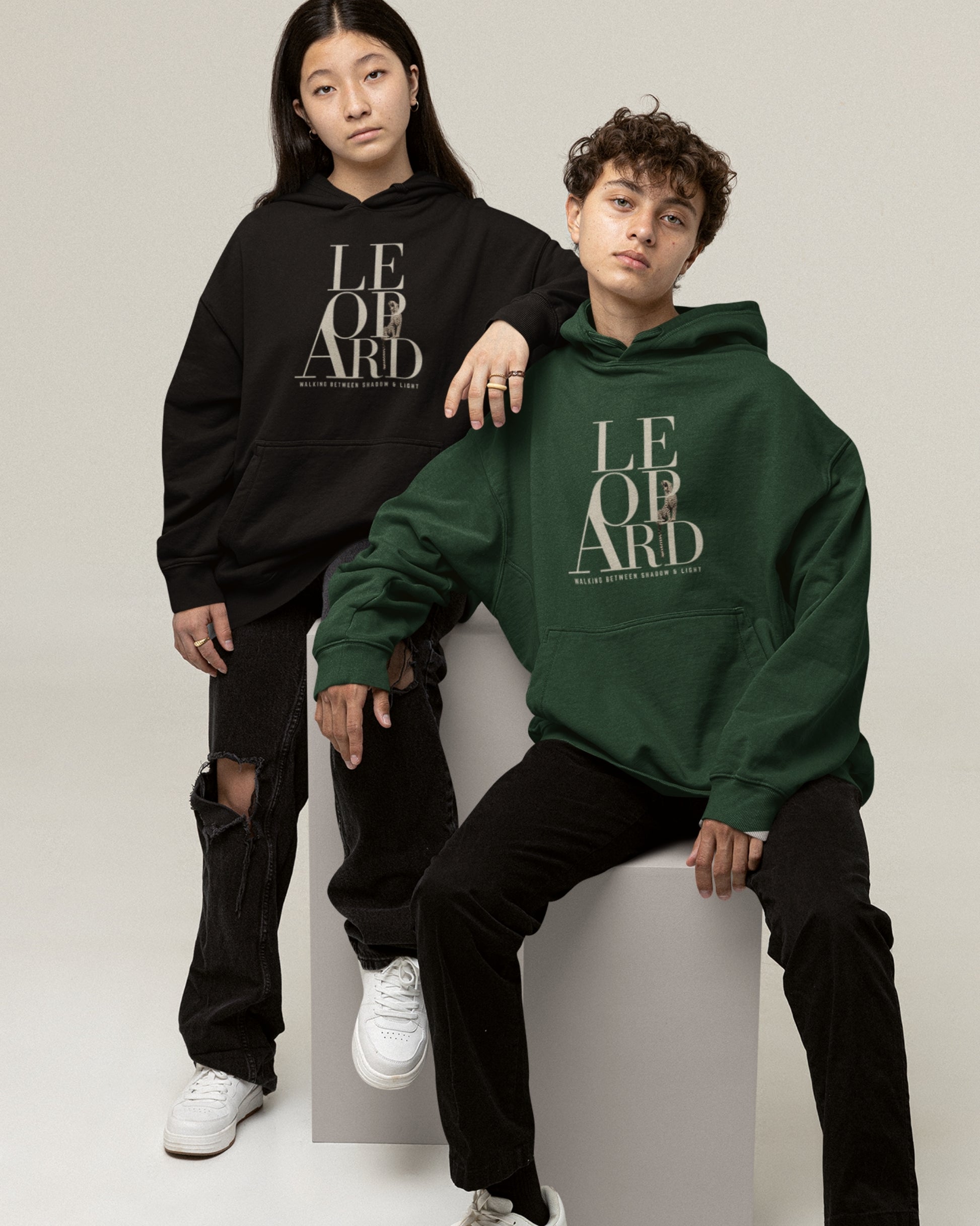 Two models posing in oversized hoodies with a leopard graphic and Walking Between Shadow and Light typography. The model in the foreground wears a dark green hoodie and black pants, sitting on a white block, while the model behind wears a black hoodie and ripped black jeans.