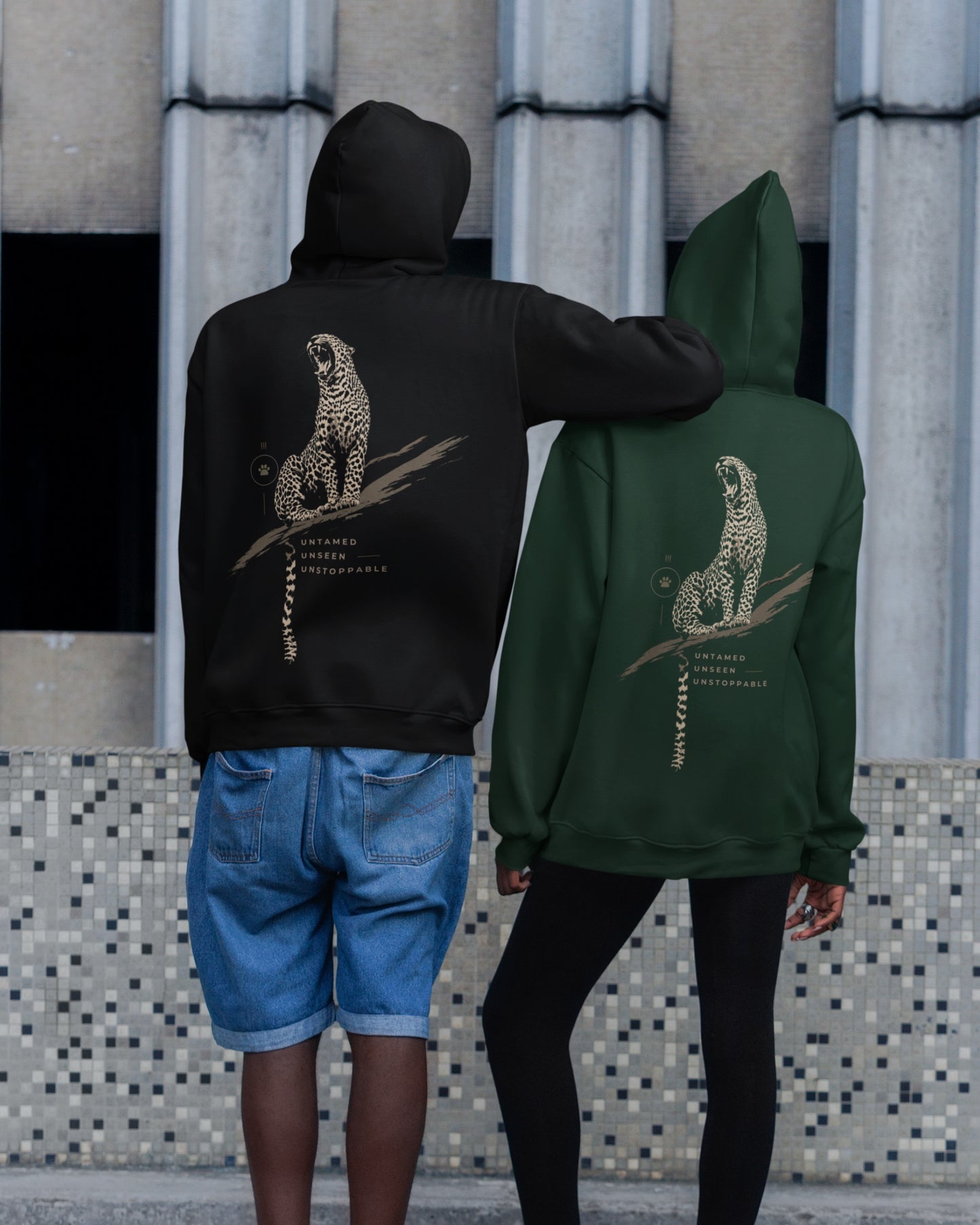 Two models from behind wearing hoodies with a large leopard graphic on the back, one in black and one in forest green, standing in front of a tiled wall.