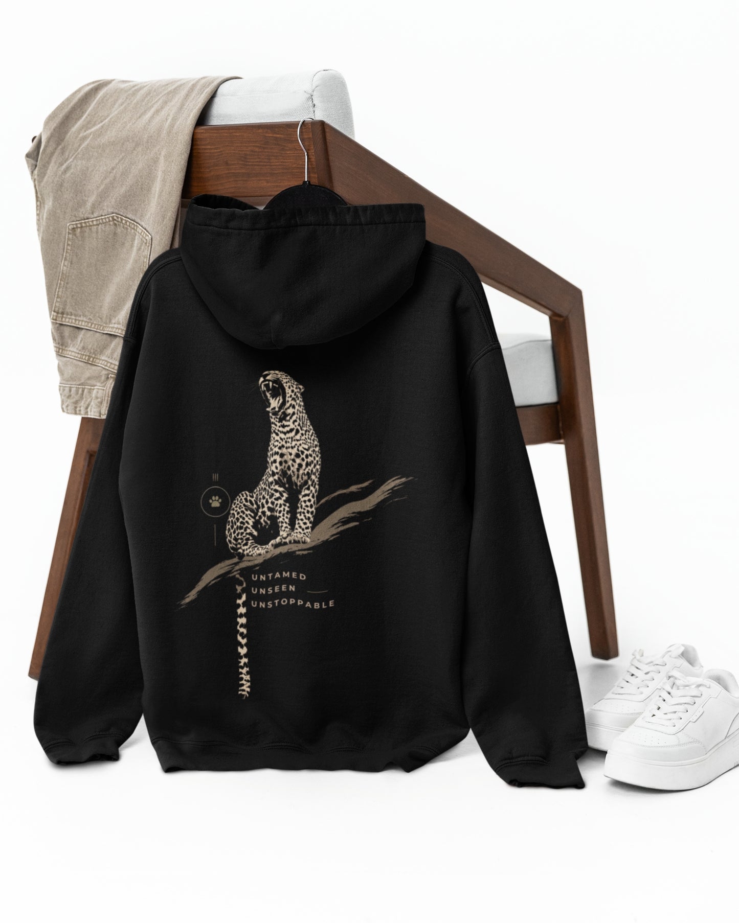 A black pullover hoodie shown from the back, styled on a wooden chair with a beige shirt draped over the backrest and white sneakers on the floor. The hoodie features a detailed graphic of a spotted big cat sitting on a branch with text underneath.