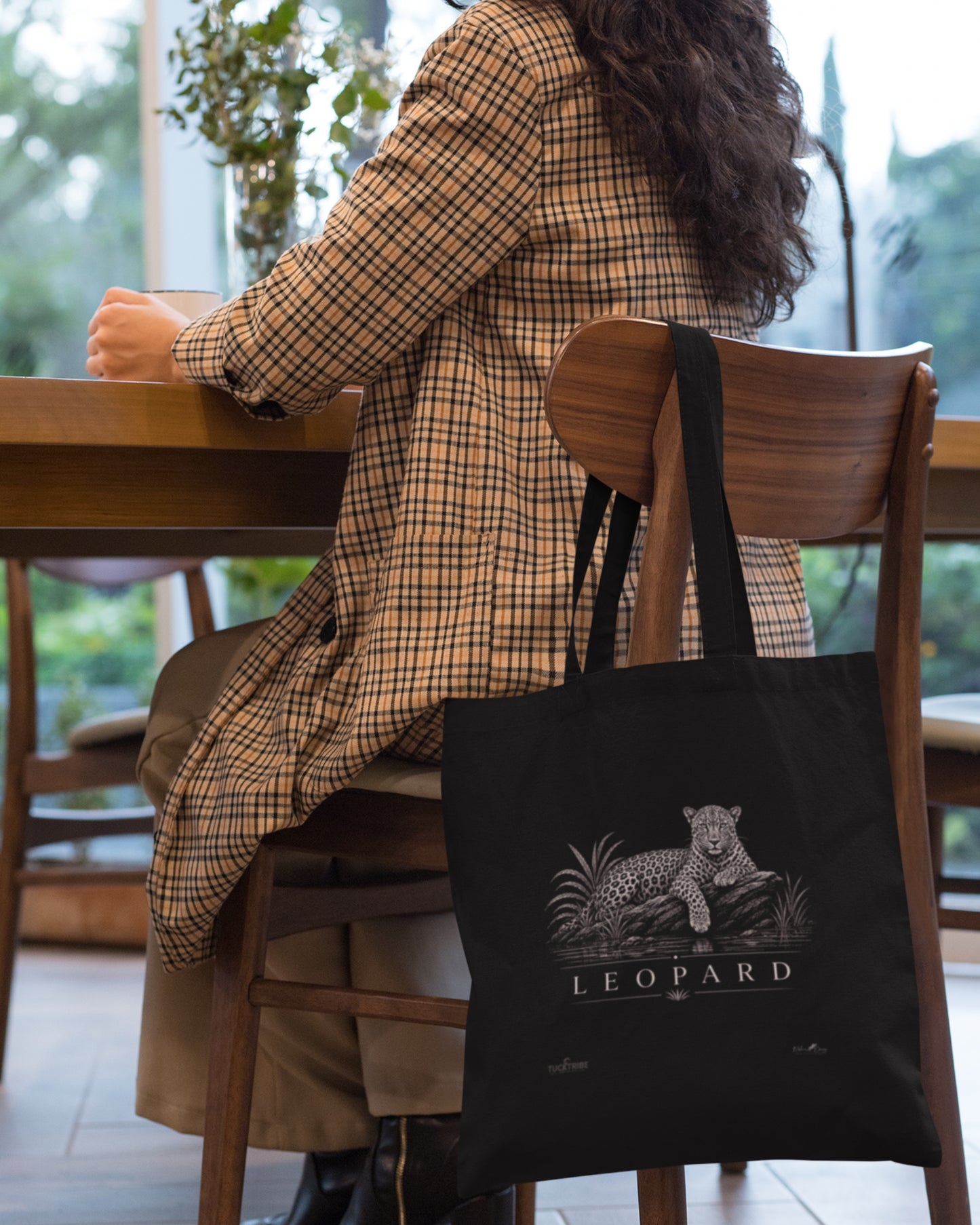 A black tucktribe canvas tote bag with a white leopard graphic and the word 'LEOPARD' sits on a wooden chair next to a woman wearing a tan and brown plaid coat, seated at a wooden table in a bright indoor setting.