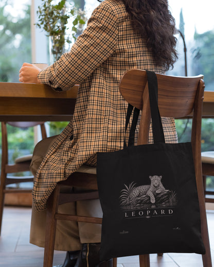 A black tucktribe canvas tote bag with a white leopard graphic and the word 'LEOPARD' sits on a wooden chair next to a woman wearing a tan and brown plaid coat, seated at a wooden table in a bright indoor setting.