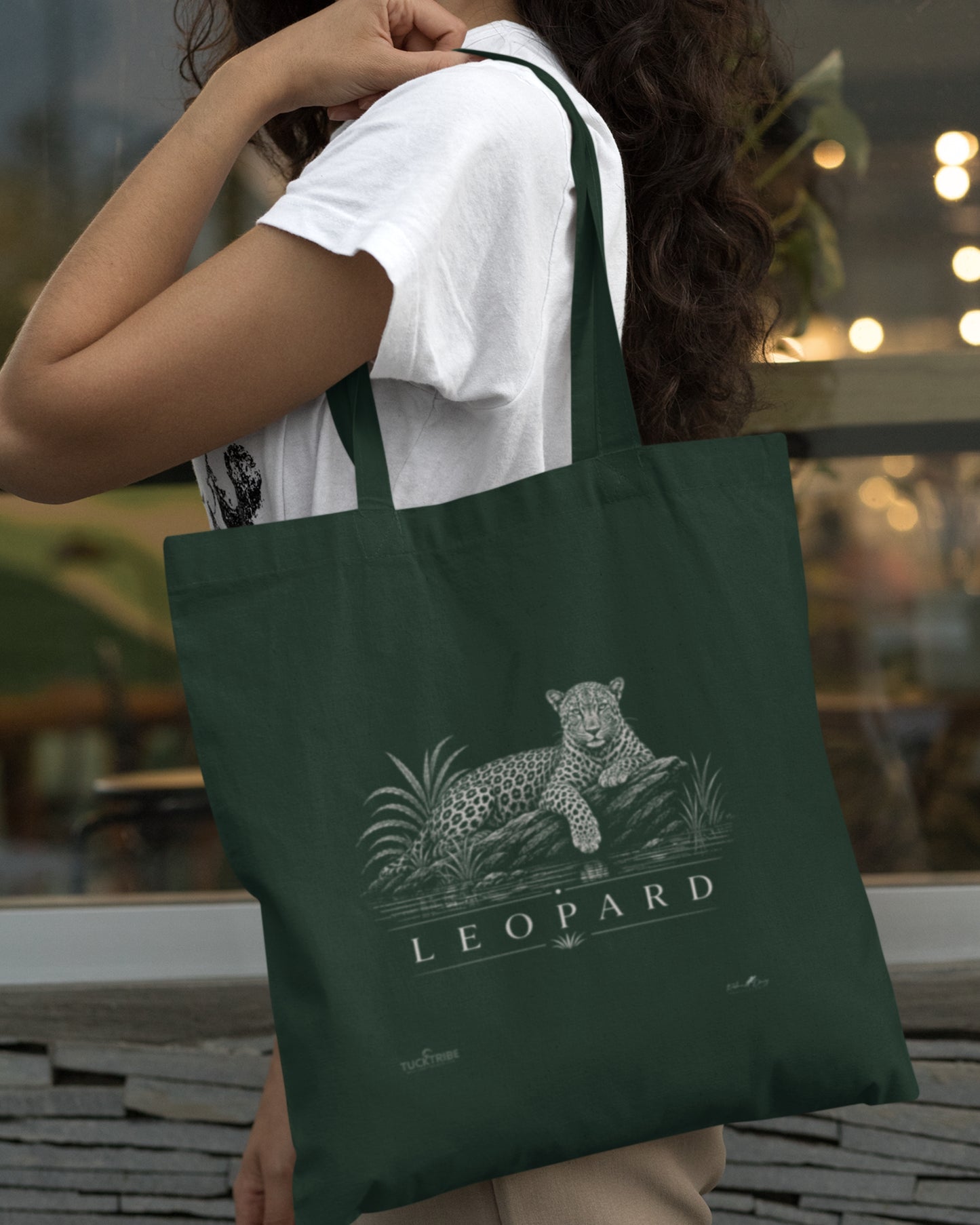 A dark green tucktribe canvas tote bag with a white leopard graphic and the word 'LEOPARD' printed on the front, carried over the shoulder of a person wearing a white t-shirt in an outdoor setting.